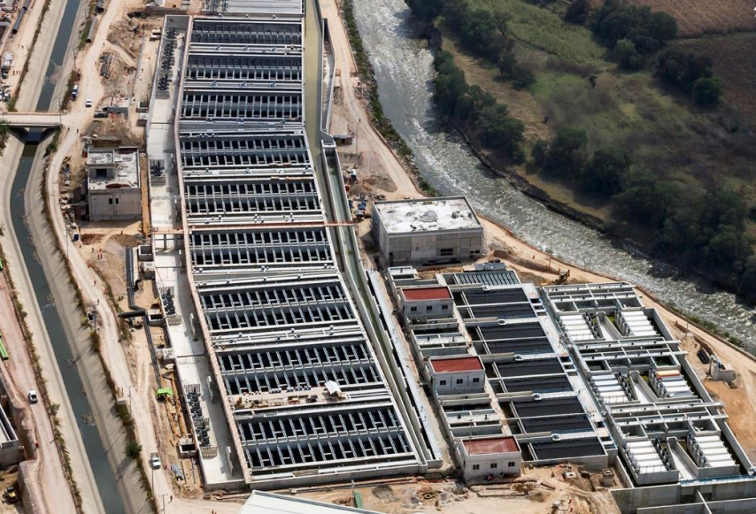 Aerial view of a large water treatment facility next to a river, featuring multiple rectangular basins, buildings, and surrounding roads. The site includes the installation of TUBEdek fills.