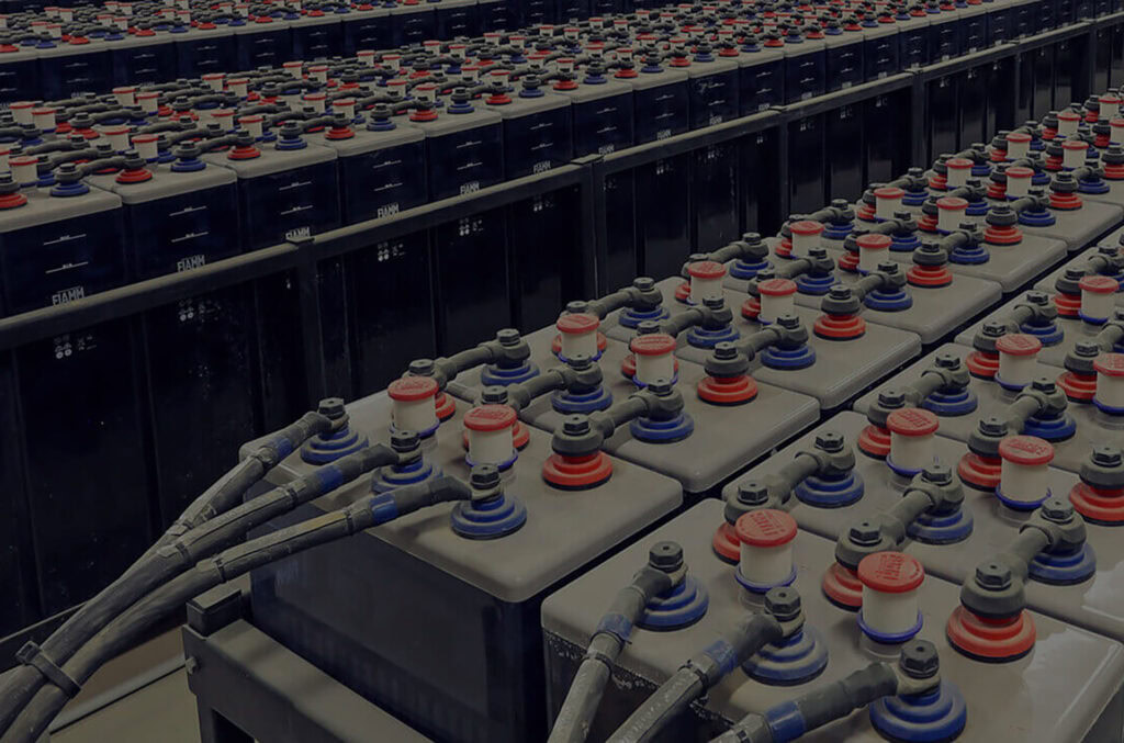 Un gran conjunto de baterías industriales están dispuestas en filas, conectadas por numerosos cables y alambres. Las baterías, con componentes rojos y azules en la parte superior y carcasas negras en la inferior, están perfectamente organizadas detrás de una placa perforada en lo que parece ser una instalación de almacenamiento o de energía.