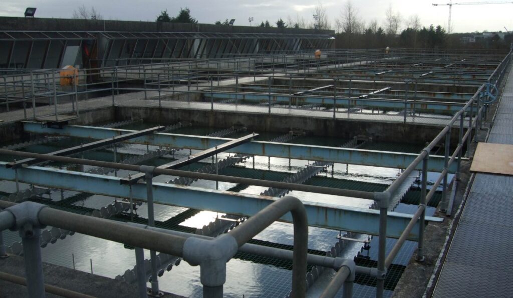 A large industrial water treatment facility with multiple rectangular tanks, interconnected by metal walkways and rails. The tanks contain water, mechanical equipment, and TUBEdek modules. The background features trees, buildings, and a crane. The sky is overcast.