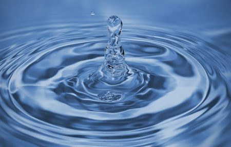 A single water droplet creates ripples on a calm, blue water surface. The droplet is caught mid-splash.