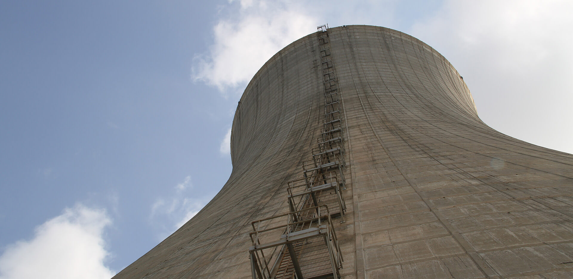 Primer plano de una gran torre de refrigeración de hormigón sobre un fondo de cielo parcialmente nublado, con una escalera metálica y una plataforma adosadas al lateral que se extienden hacia la parte superior de la estructura.