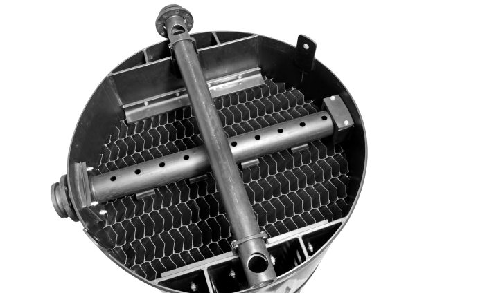 Close-up of a cylindrical industrial component resembling a sedimentation tank with a grid-like structure inside. Two horizontal pipes with multiple holes run through the center of the cylinder, and there are several connectors and brackets attached to the edges.
