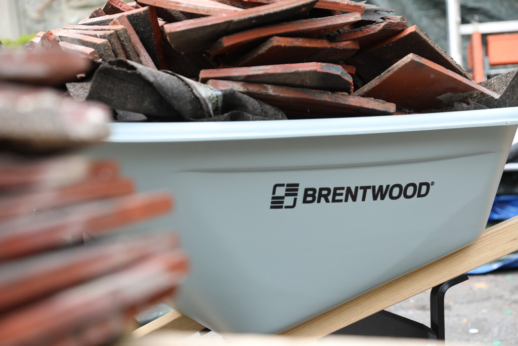 A white Brentwood wheelbarrow, is filled with broken bricks and debris. More bricks blur into the foreground, while a cluttered workspace provides the backdrop.