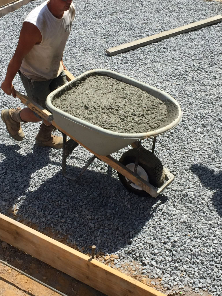 A person pushes a wheelbarrow filled with wet concrete over a bed of gravel at a construction site. They are wearing a sleeveless shirt, shorts, and work boots. Wooden planks and other landscaping equipment are visible in the background.