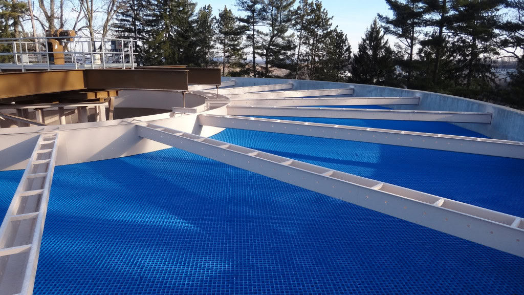 A circular industrial structure with angled beams supporting a blue grid on the ground, part of a water treatment or wastewater filtration system. The background features tall trees under a clear sky.