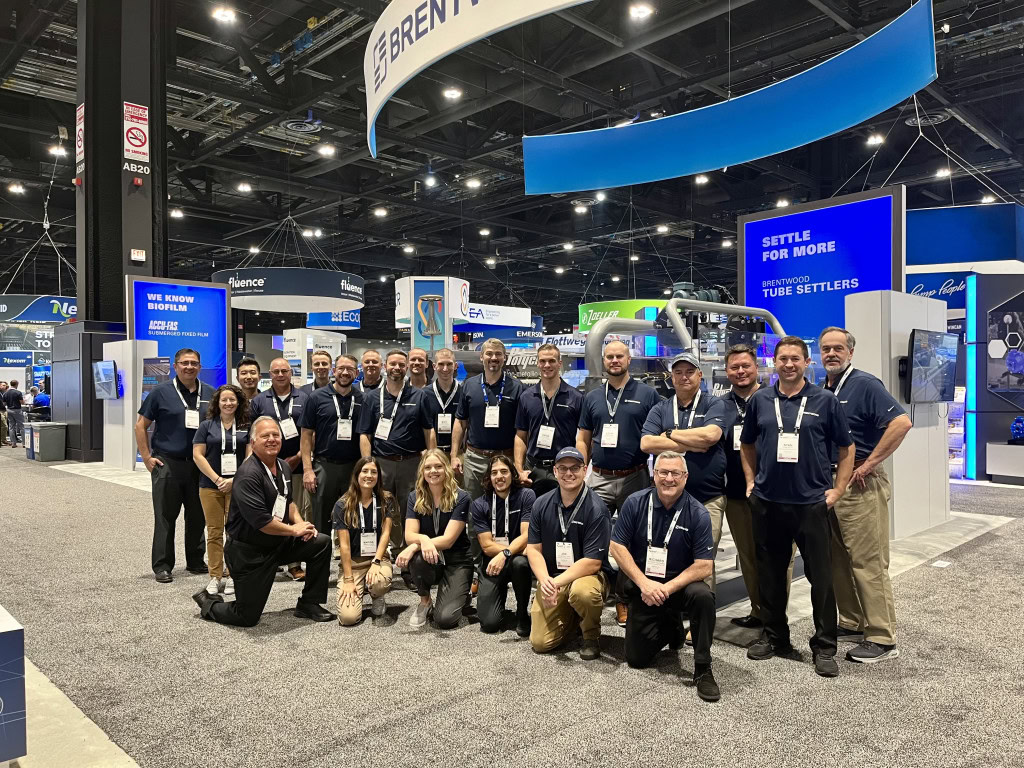 A large group of people wearing navy blue Brentwood shirts with name tags pose for a group photo at an exhibition booth labeled "Brentwood Wastewater." The background features other exhibition booths with bright displays and high ceilings in a convention center.