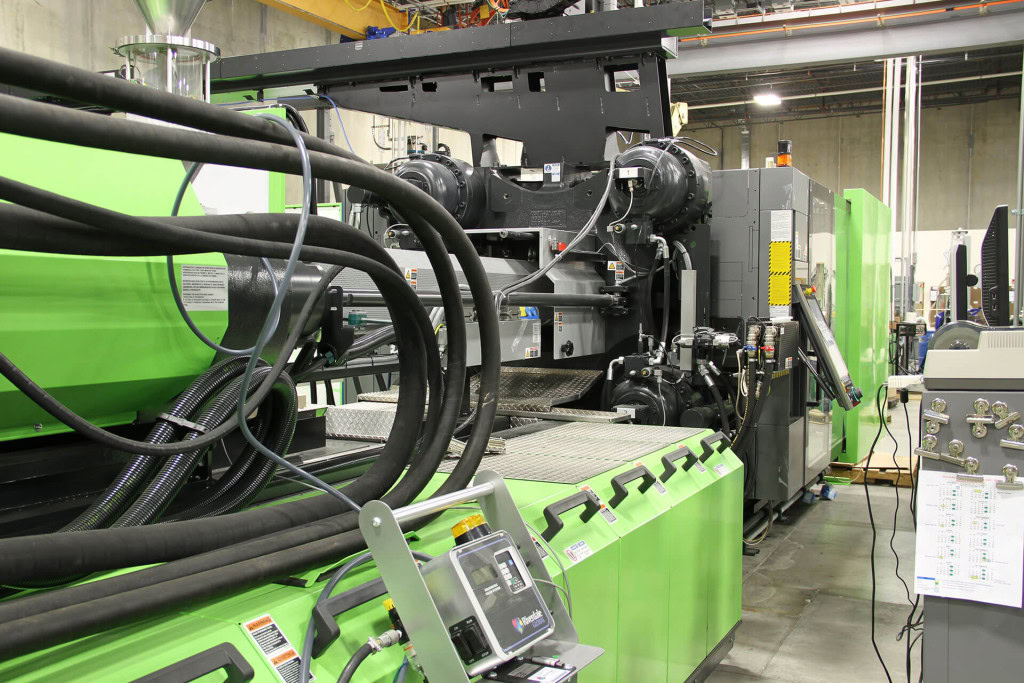 A large industrial injection molding machine in a factory setting. The primarily green machine, with black hoses and other machinery components, is surrounded by a clean and organized environment.