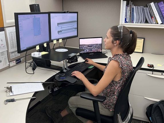 A person is sitting at an office desk, working on two computer monitors displaying technical designs. A laptop sits beside the monitors. The desk has various papers, a clipboard, and office supplies.