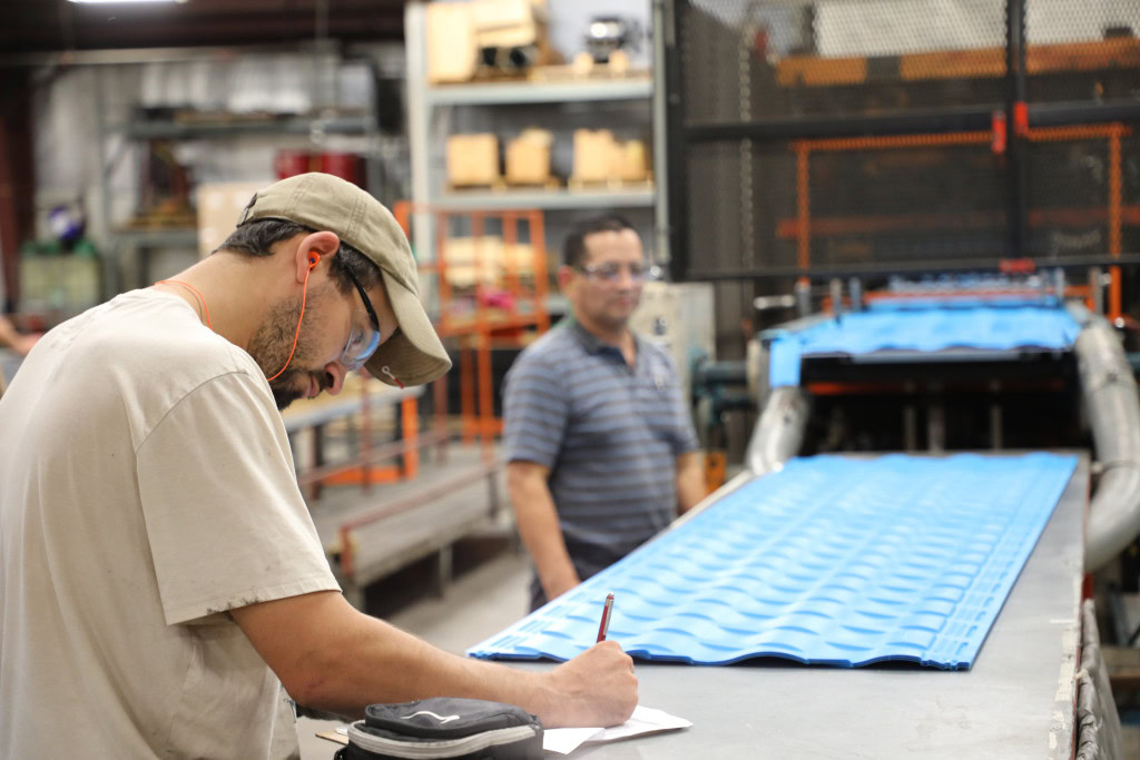 A man in a beige shirt and cap is writing on a clipboard in a factory setting. In the background, another man in a striped shirt stands near a machine producing blue, corrugated panels.