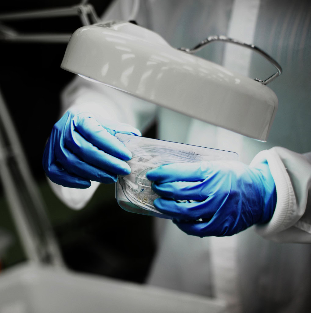 una persona con guantes azules y bata blanca de protección, trabajando bajo una lámpara brillante, sosteniendo un recipiente de plástico transparente