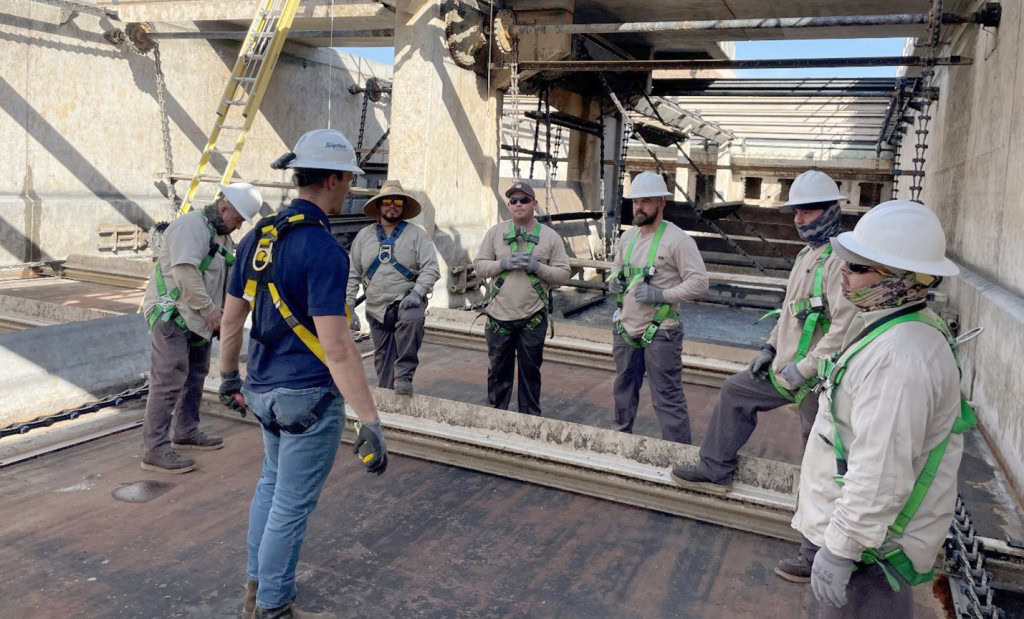 A group of six construction workers wearing safety gear, including hard hats and harnesses, stand in a semi-circle around a supervisor on a construction site. The supervisor is wearing a navy shirt and helmet.