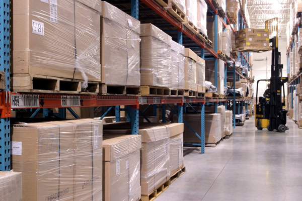 A warehouse scene showing rows of shelves filled with large, wrapped boxes and pallets. A forklift is visible in the aisle, lifting a pallet.