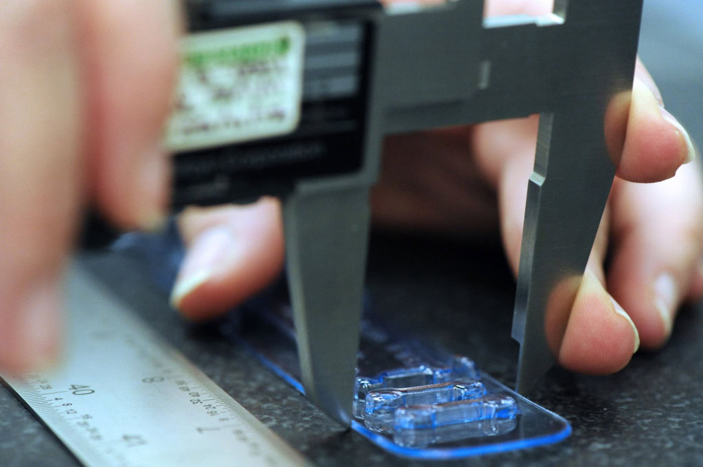 Close-up image of a person using a digital caliper to measure the thickness of a clear plastic object. A metal ruler lies next to the object on a dark surface.