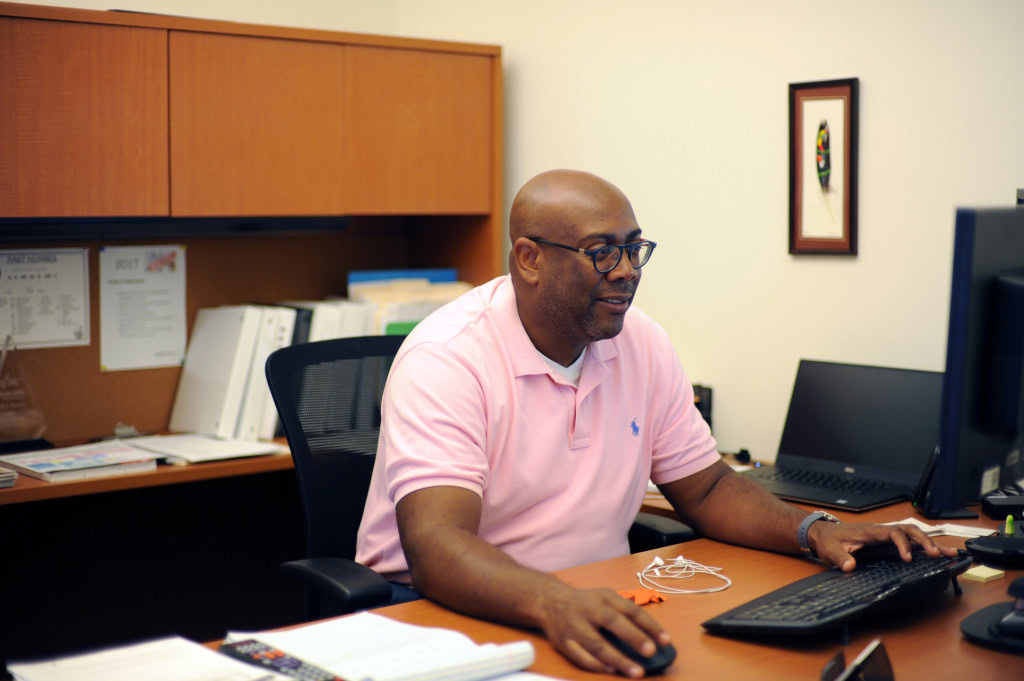 A man wearing glasses and a pink polo shirt sits at a desk working on a computer. The desk has various office supplies and documents. In the background, there are shelves with binders.