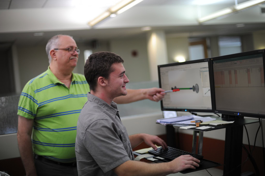 Two men, one older with glasses and a striped polo shirt and one younger in a gray shirt, work at a standing desk with dual monitors. The younger man types while the older man points to a 3D model on one of the screens.