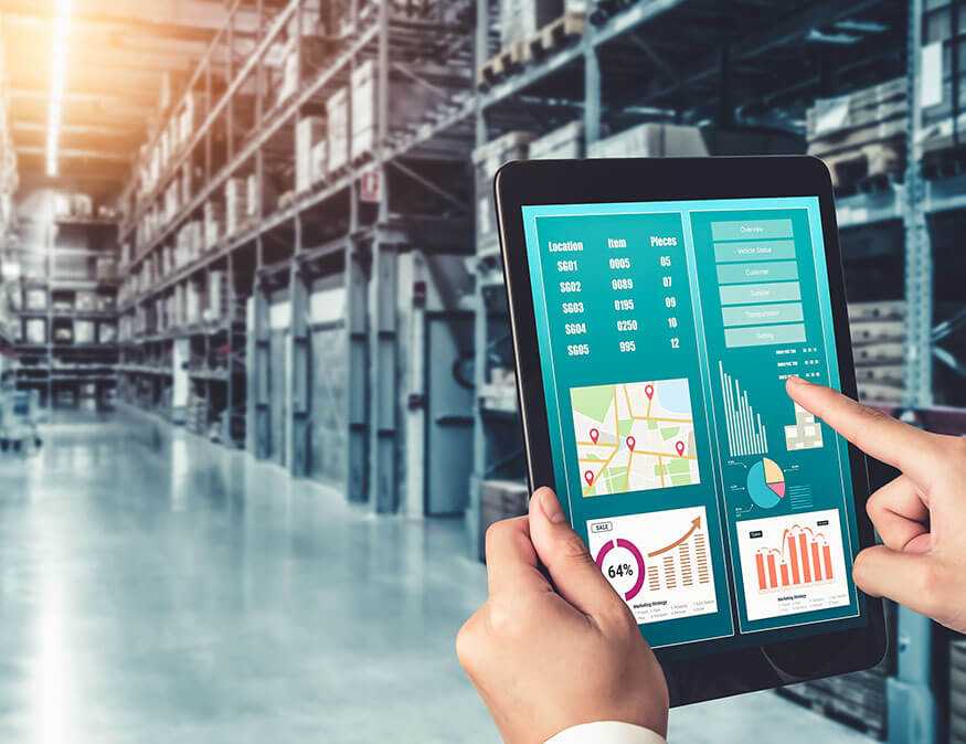 A person holds a tablet displaying logistics data, including maps, bar charts, and graphs while standing in a large, organized warehouse with shelves stocked with various items.