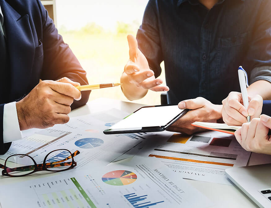 Three individuals engage in a business meeting, discussing financial documents on a table. One points at a tablet screen, another holds a pen, and various charts and graphs are visible.