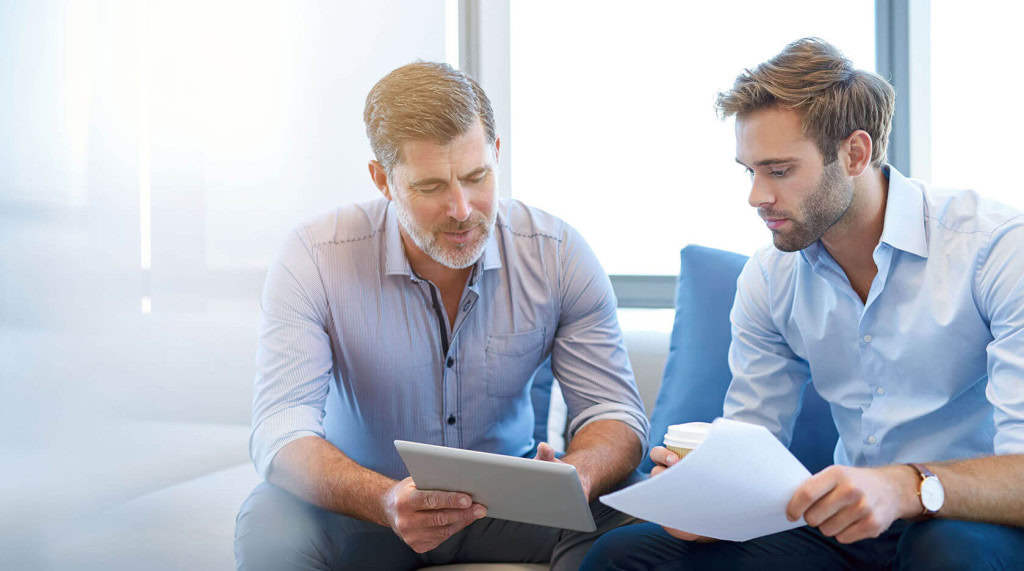 Two men are sitting on a couch in a bright office, engaged in a conversation. The older man on the left is holding a tablet and showing something to the younger man on the right, who is holding a piece of paper and a cup.