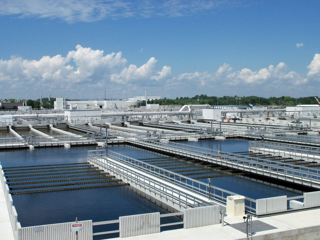 A large water treatment facility with multiple rectangular pools containing water. Walkways and railings run alongside and between the pools. Buildings and machinery are visible in the background under a bright blue sky with scattered clouds.