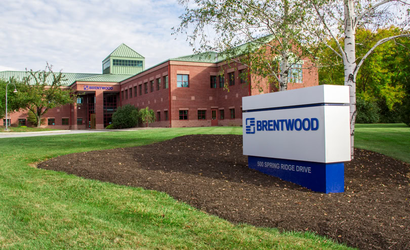 A red-brick office building with a green metal roof labeled "Brentwood." In front of the building, there is a white and blue sign reading "Brentwood" and "500 Spring Ridge Drive."