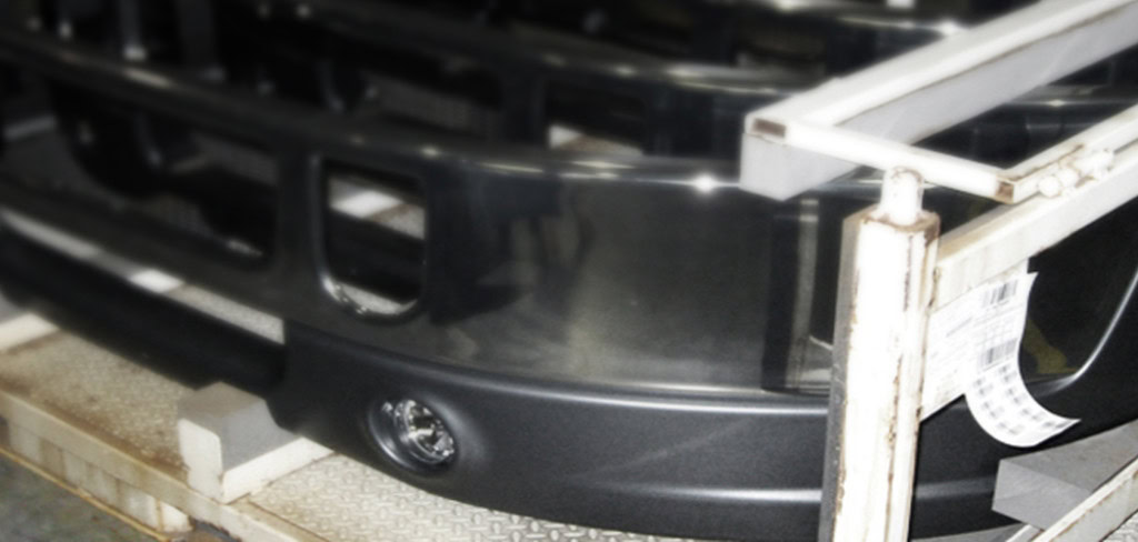 A close-up image of a vehicle's front bumper being manufactured showcases the meticulous craftsmanship involved. The black, partially wrapped bumper rests on a metal support frame.