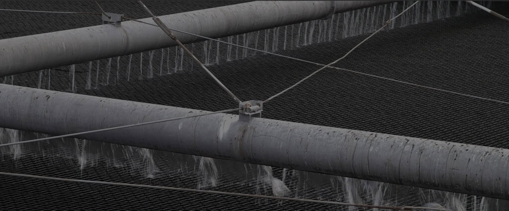 Primer plano de grandes tuberías industriales con agua cayendo en cascada a través de una estructura de malla metálica. Las tuberías, que transportan aguas residuales, se apoyan en una red de cables, creando una disposición compleja pero organizada.