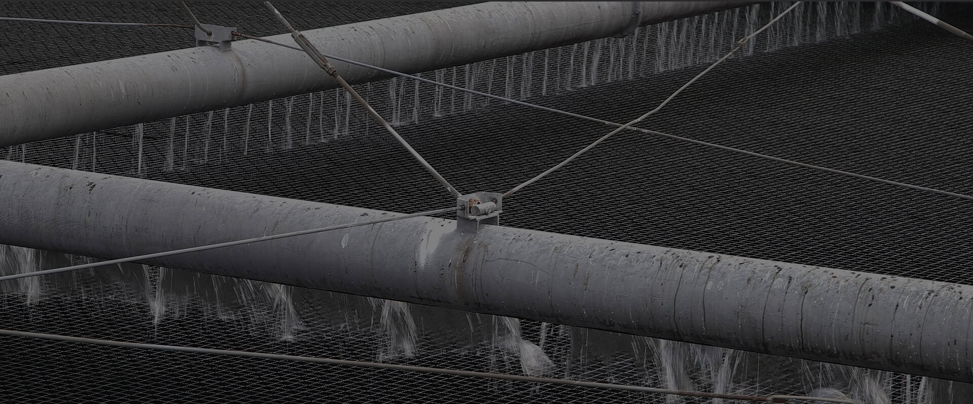 Close-up of large industrial pipes with water cascading through a metal mesh structure. The pipes, carrying wastewater, are supported by a network of cables, creating a complex yet organized arrangement.