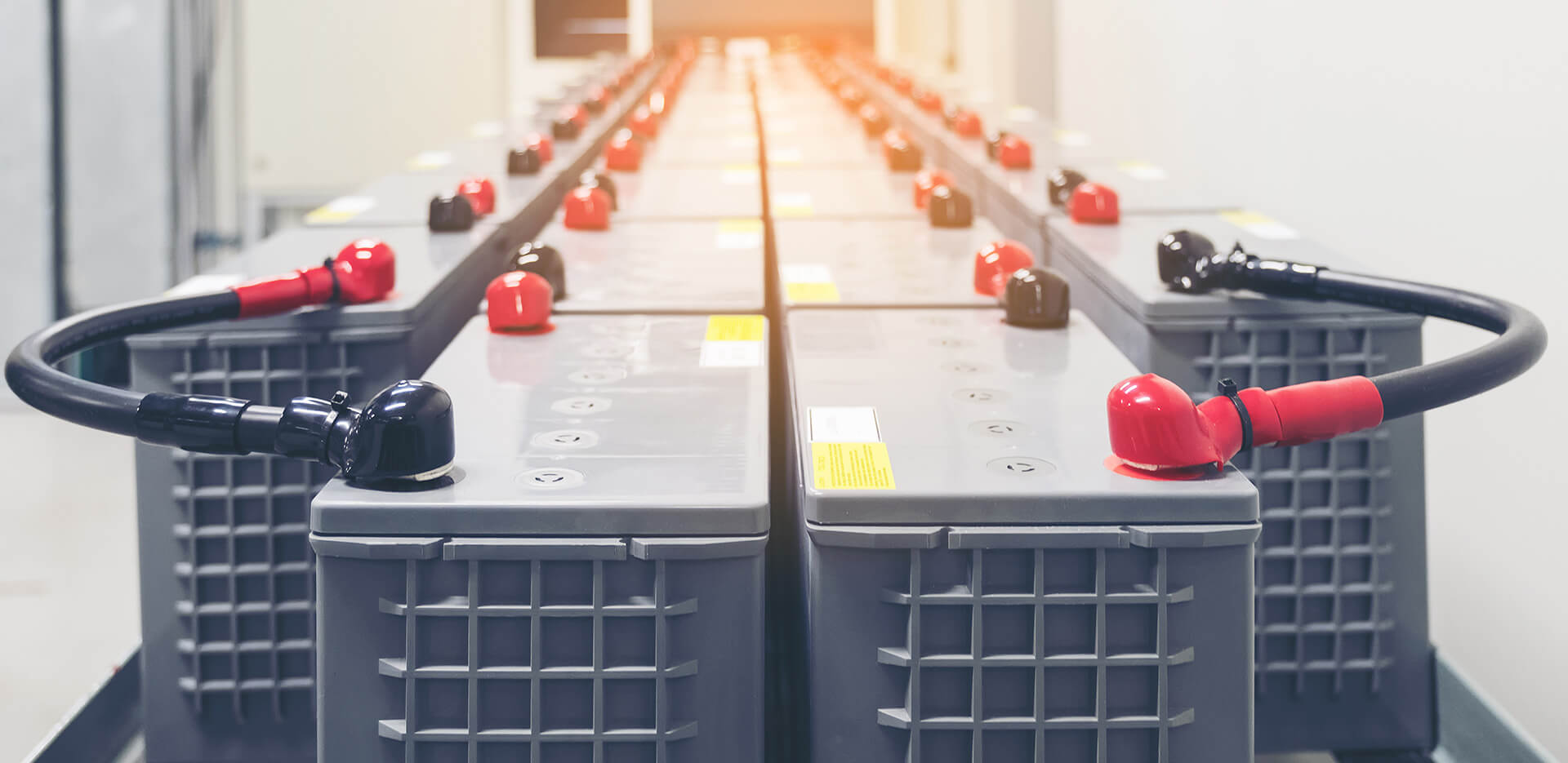 Una fila de baterías industriales dispuestas simétricamente en una habitación muy iluminada. Cada batería está provista de bornes rojos y negros, con cables conectados.