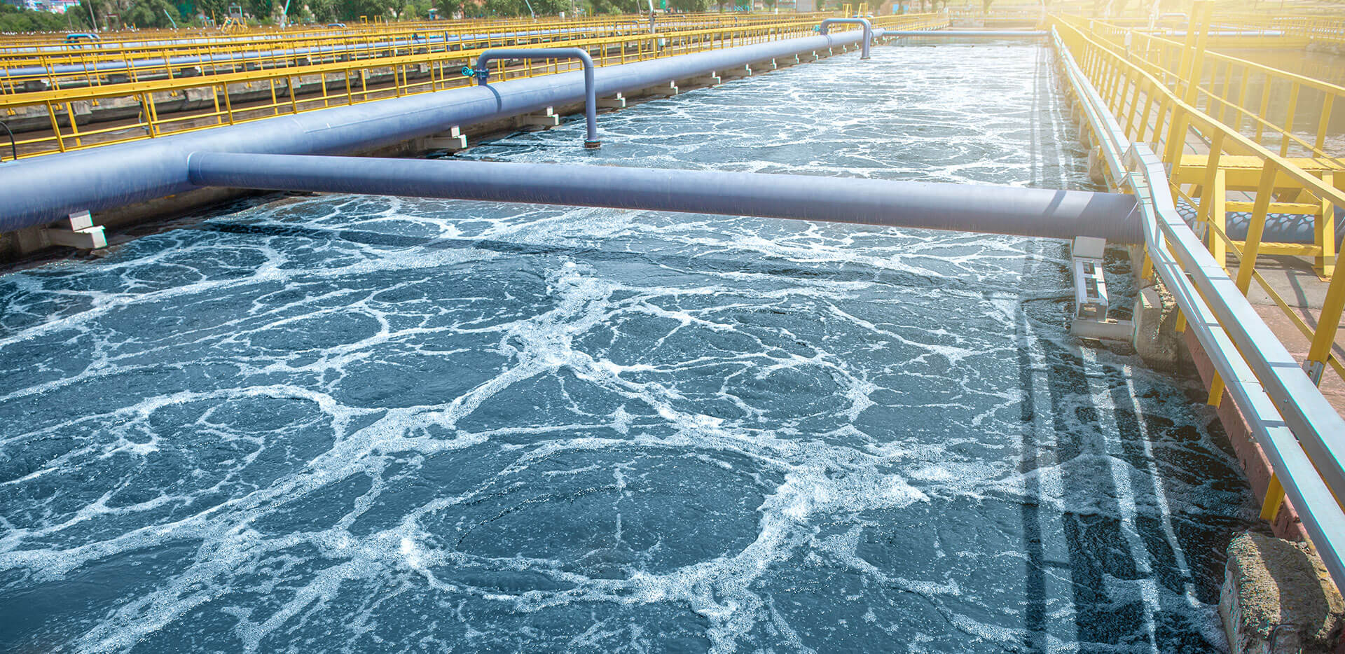 Una gran instalación industrial de tratamiento de aguas con agua aireada en un depósito largo y rectangular. Múltiples tuberías y barandillas amarillas discurren junto al agua.