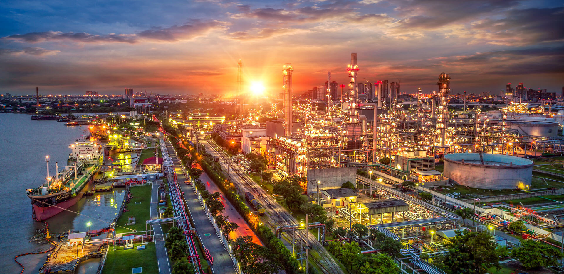 Junto a un río, bajo un cielo al atardecer, se ve un extenso complejo industrial con numerosas estructuras y tanques iluminados.